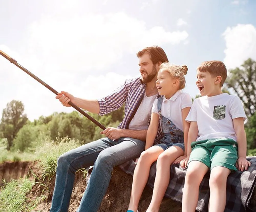 A man and two children are sitting by a riverbank smiling and taking a selfie on a sunny day