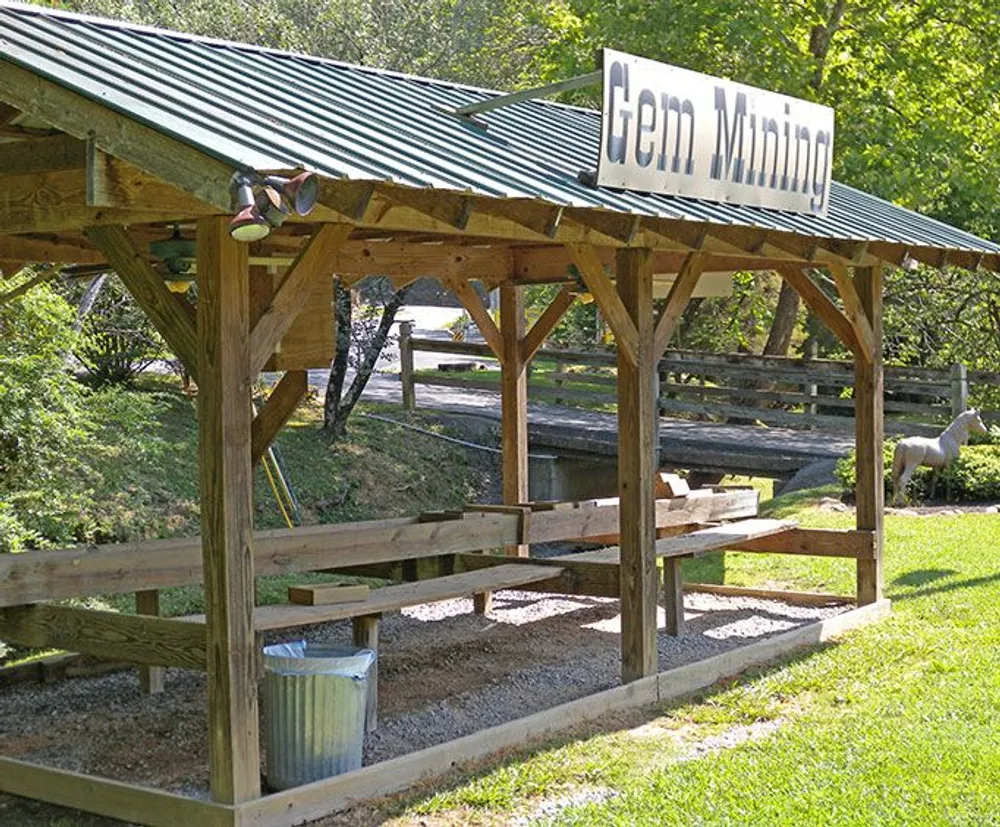 The image shows an outdoor gem mining station with a long wooden sluice structure under a roof and a sign that reads Gem Mining