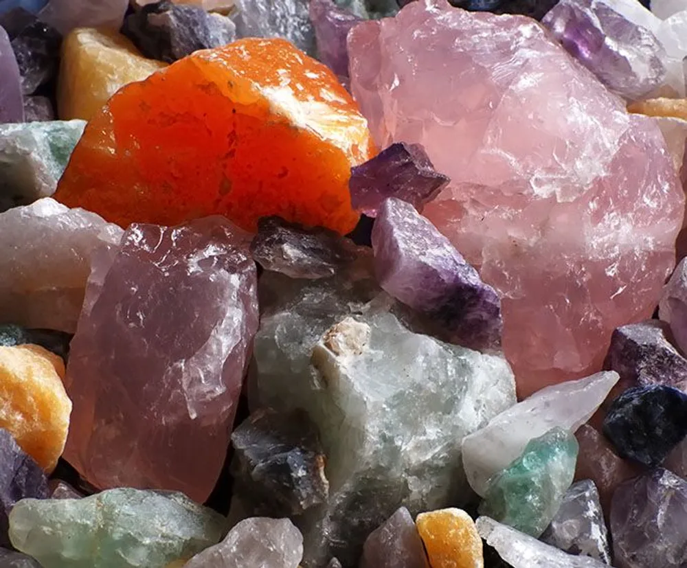 The image shows a close-up of a colorful assortment of rough gemstones and mineral crystals with various sizes and hues