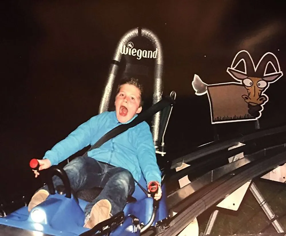A child appears thrilled and possibly a bit scared riding a roller coaster at night with an illustrated goat character displayed beside the track