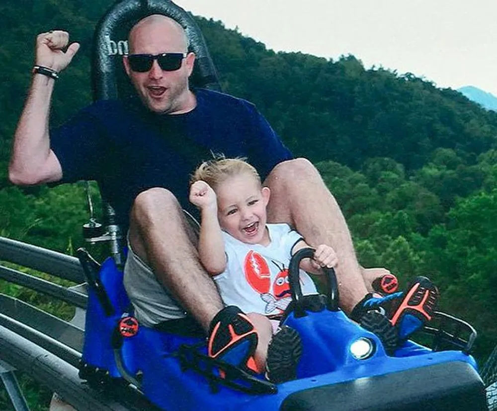 An adult and a child are raising their arms in excitement while riding a blue alpine coaster amidst a lush green landscape