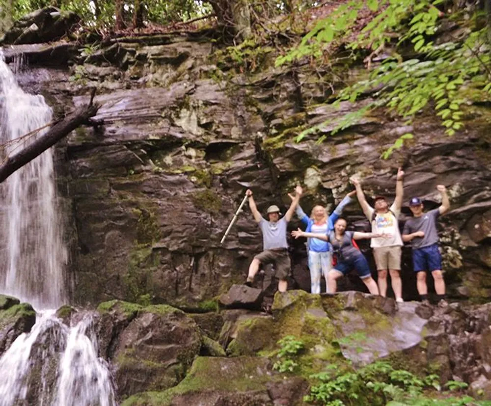 A group of five people is posing joyfully in front of a waterfall on a rocky ledge with some holding hiking sticks and all raising their arms or sticks in a celebratory manner