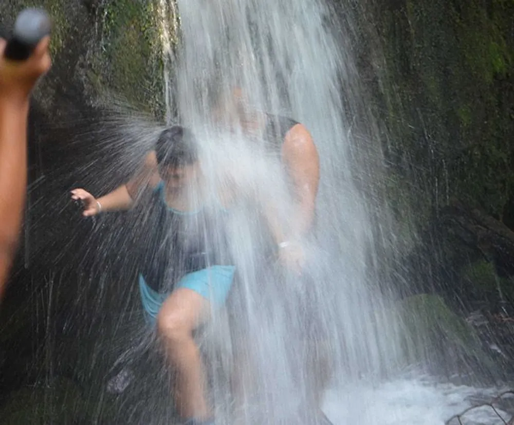 A person is taking a shower under a natural waterfall appearing to enjoy the experience amidst a misty woodland setting