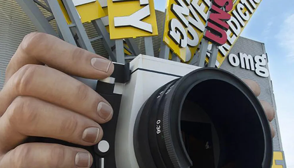 The image is a clever perspective illusion of a giant hand holding a camera with the background signage appearing to extend from the camera as if it were a snapshot being taken