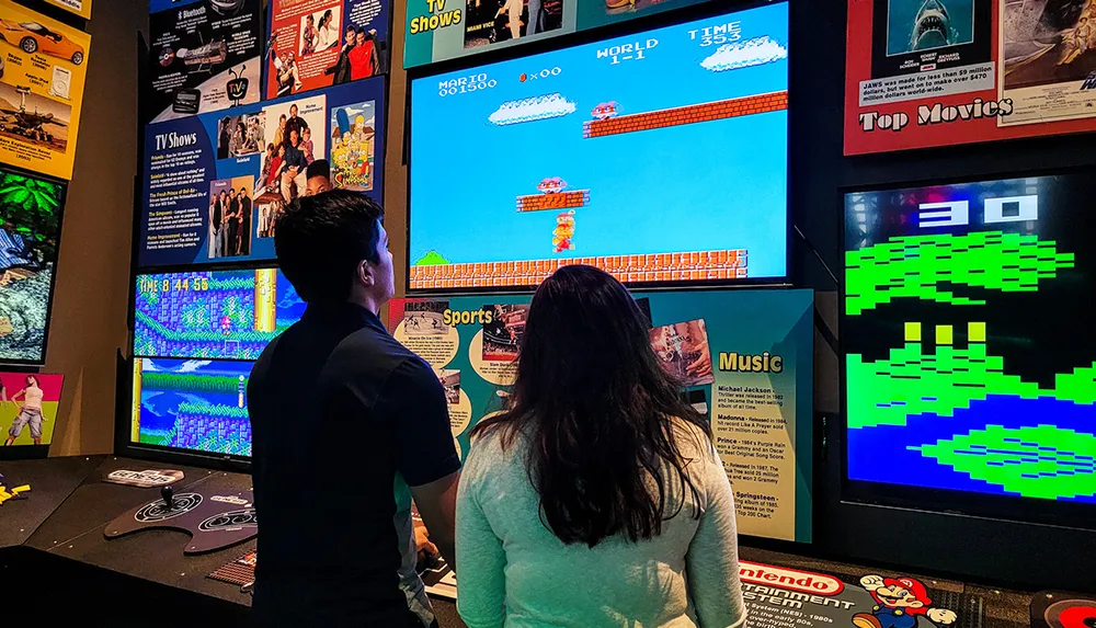 Two people are playing a classic Super Mario Bros video game on a large screen in a room that also features various posters likely related to pop culture and media history