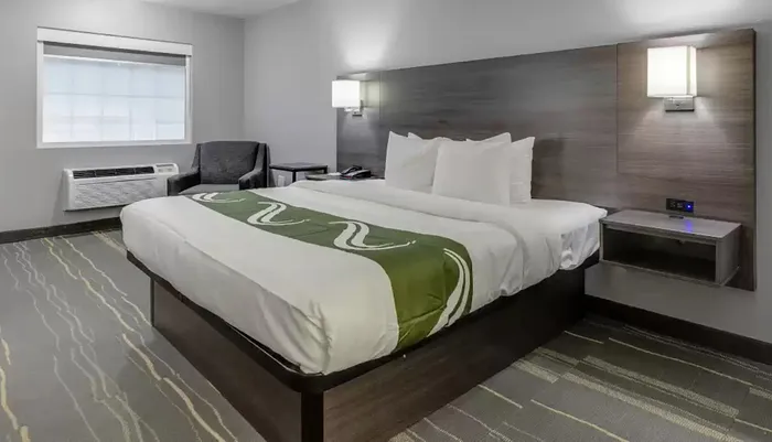 The image shows a modern hotel room with a neatly made king-size bed a patterned carpet a wall-mounted TV a side chair an air conditioner and a simple decor