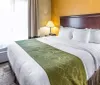 The image shows a neatly prepared hotel room with a large bed crisp white linens and a green decorative runner at the foot of the bed accompanied by wooden furniture and warm lighting that creates a welcoming atmosphere
