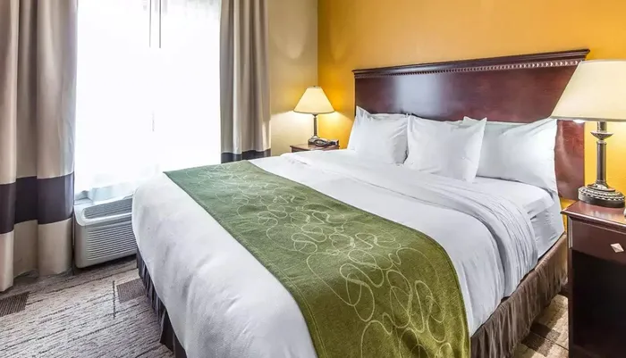 The image shows a neatly prepared hotel room with a large bed crisp white linens and a green decorative runner at the foot of the bed accompanied by wooden furniture and warm lighting that creates a welcoming atmosphere