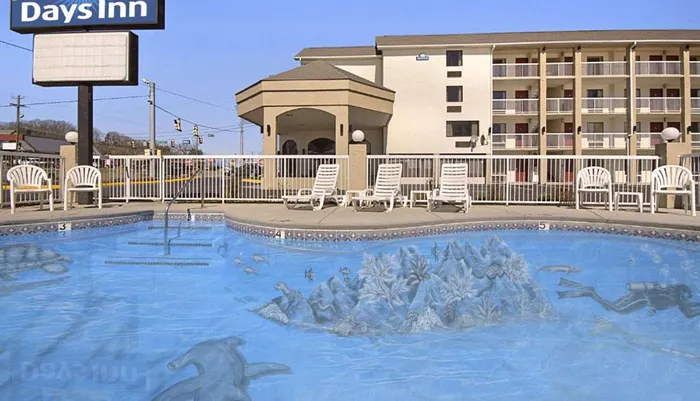 This image depicts an outdoor swimming pool with a decorative underwater mural surrounded by white chairs and a Days Inn hotel in the background