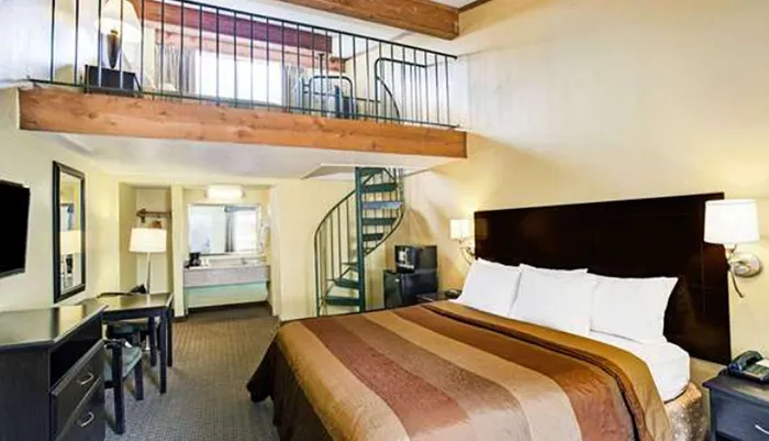 The image shows a hotel room with a large bed a work desk a spiral staircase leading to a loft area and a flat-screen TV on the wall