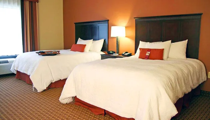 The image shows a hotel room with two neatly made queen-sized beds a nightstand with a lamp and a warm color scheme