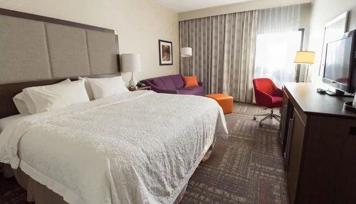 This image depicts a neatly arranged hotel room with a large bed a purple sofa a small orange ottoman and a work desk with a television and a red chair near the window