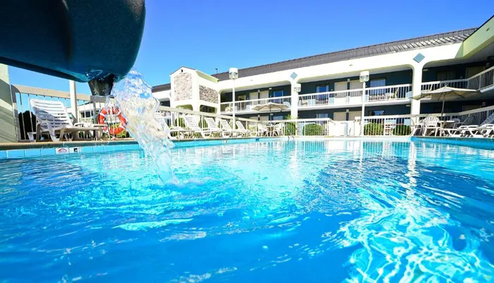 The image shows a clear blue swimming pool with water flowing into it from a fountain framed by a hotel with multiple balustraded balconies and loungers under a bright blue sky