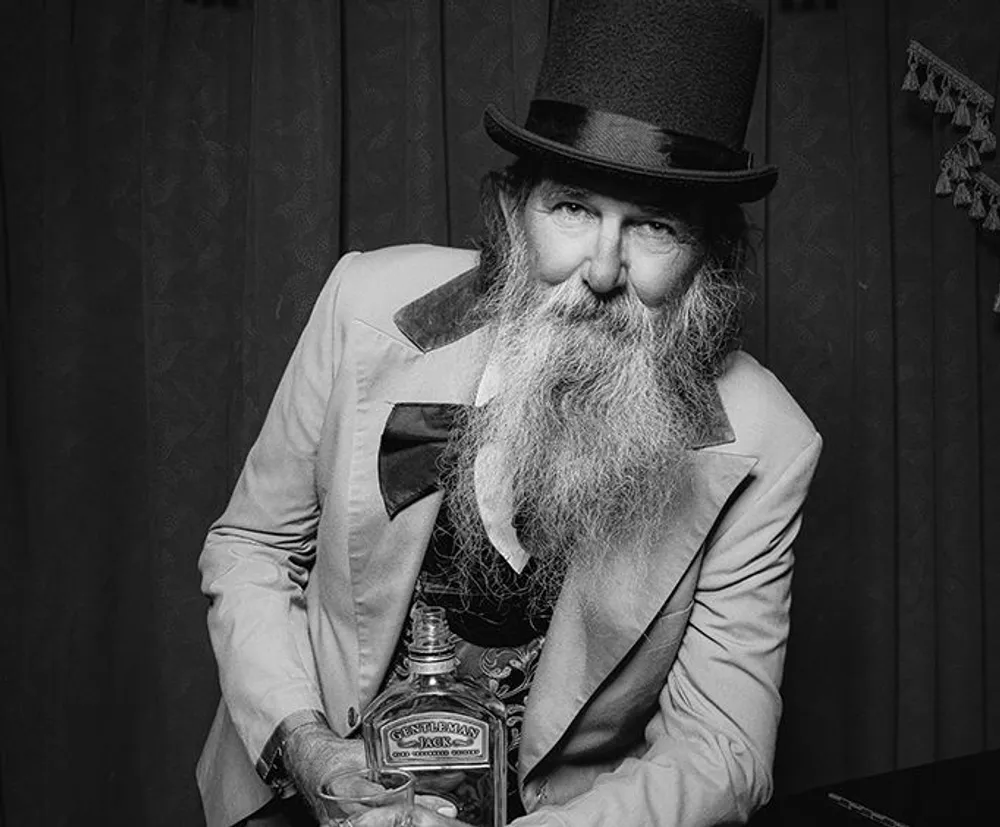 A bearded man in a top hat and a vintage suit holds a bottle and a glass giving off a classic old-timey vibe