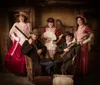 Five individuals are posed in an old-time western setting with the women dressed in period costumes with hats and holding fans while the men seated with rifles are attired in cowboy hats and dusters