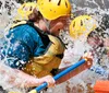 A group of people wearing life jackets and helmets are joyfully white water rafting splashing through a rapid