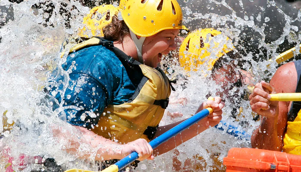 Two people in safety helmets and life vests are vigorously paddling through splashing water likely engaged in a white-water rafting adventure