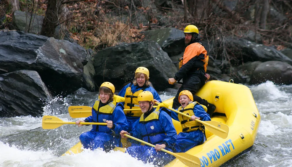 A group of people wearing yellow helmets and blue life vests enthusiastically paddle a yellow raft down a frothy river