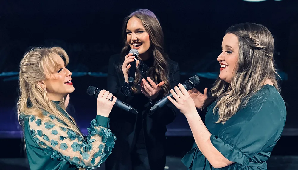 Three women are joyfully singing into microphones on a stage