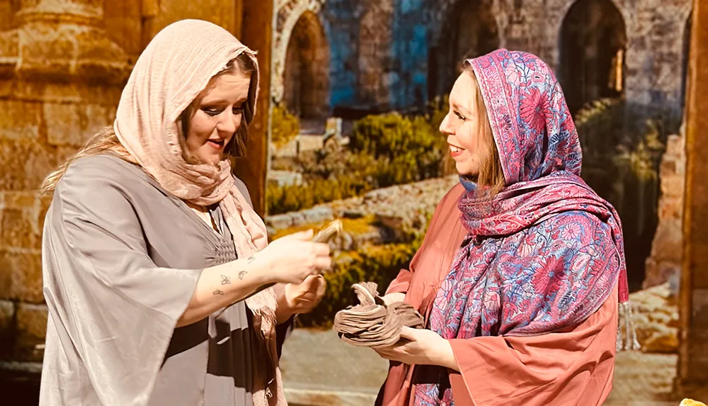 Two women one passing a piece of fabric to the other are engaged in a friendly exchange with historical or cultural context given their traditional headscarves and backdrop suggesting an ancient setting