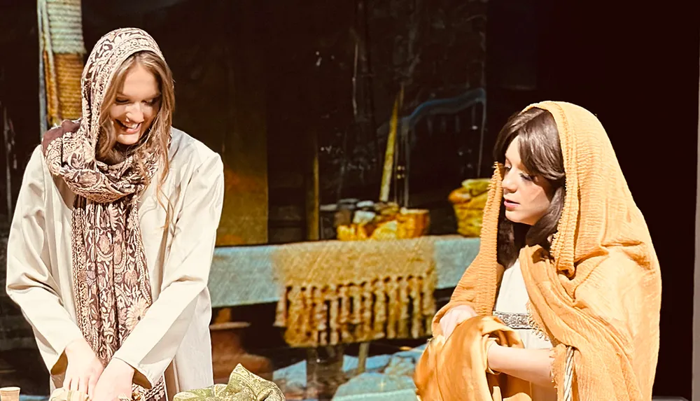 Two women dressed in traditional attire appear engaged in an interactive moment one smiling and the other with a contemplative expression in a setting with rustic and cultural elements