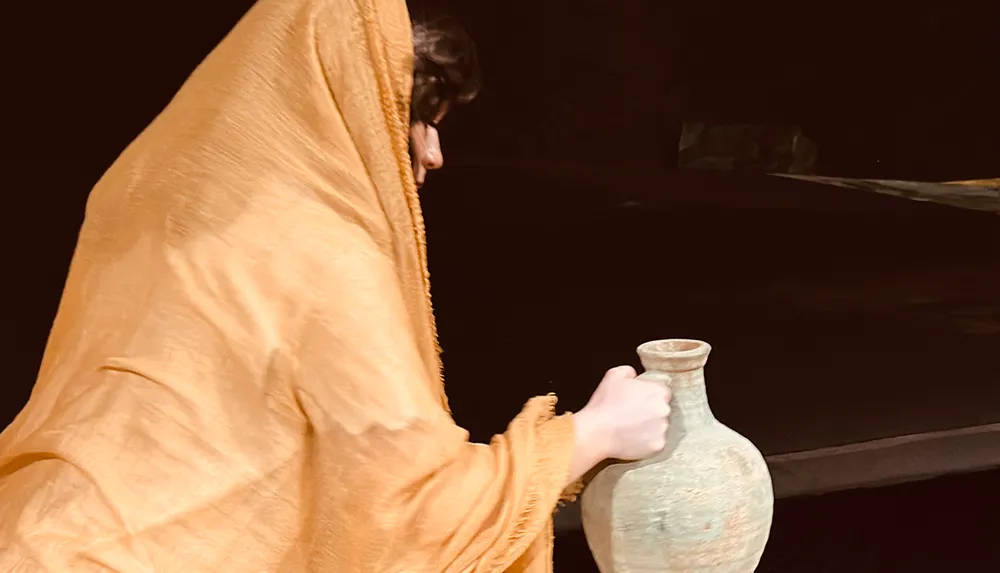 A person cloaked in a golden-yellow garment is holding an ancient-looking pottery vase