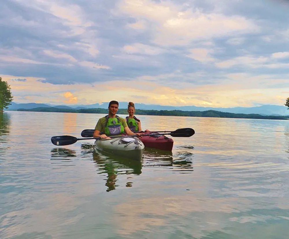 Two people are enjoying a kayak adventure on a serene lake with a picturesque backdrop of mountains and a cloud-filled sky