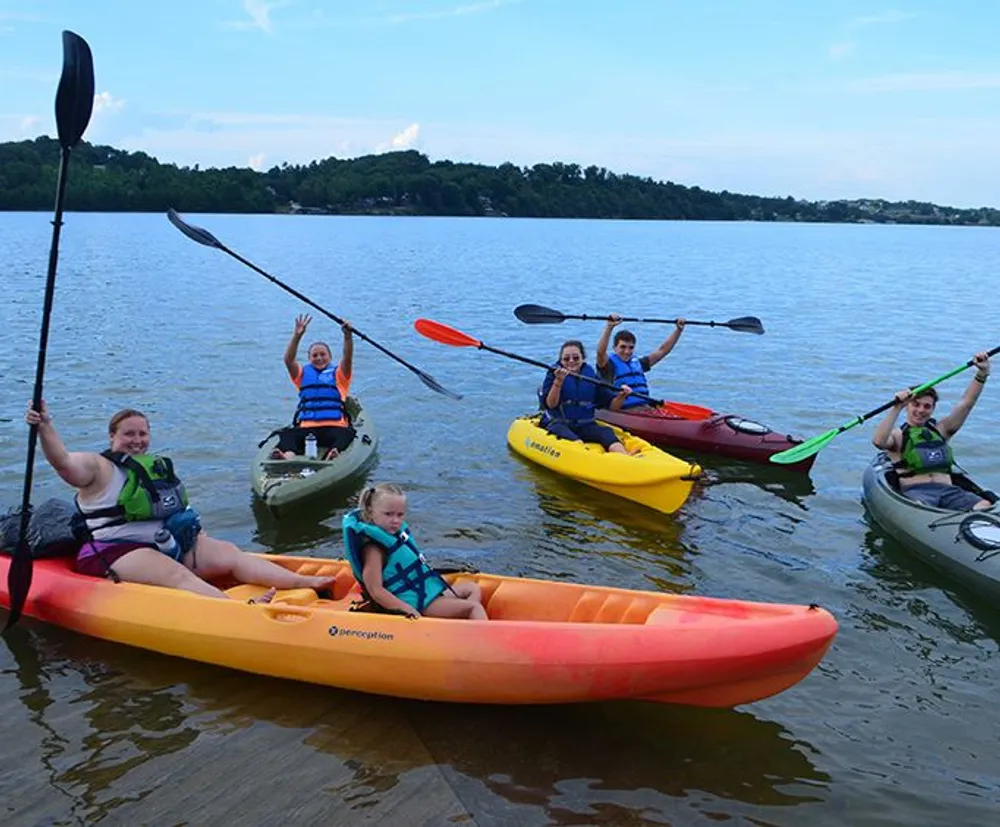 A group of smiling people are seated in colorful kayaks holding paddles and appear ready for a paddling adventure on a calm lake
