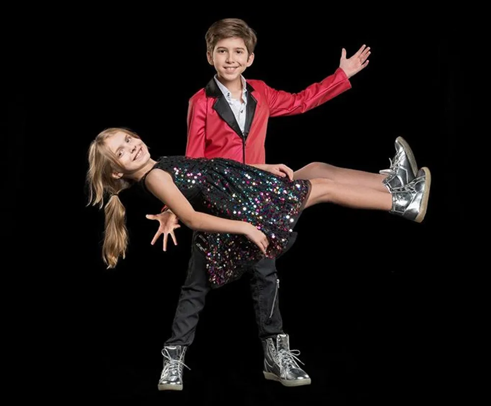 A boy in a red jacket is smiling and holding a girl in a sequined dress who is playfully posing as if flying both against a black background