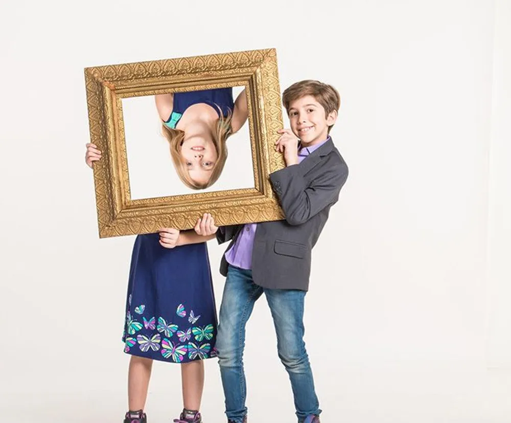 Two children are playfully posing with a golden picture frame with the girl upside down inside the frame and the boy holding it on one side both smiling