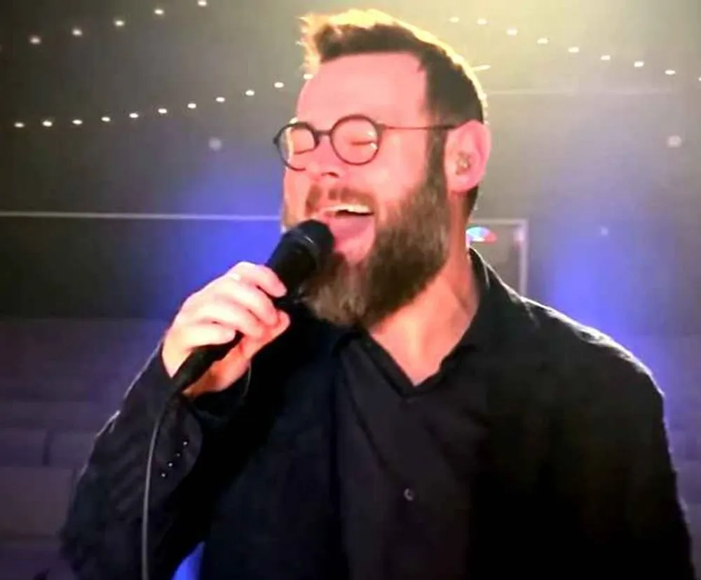 A bearded man with round glasses is passionately singing into a microphone with a blurred background that suggests a stage with lights