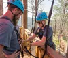 Two people wearing helmets and harnesses are happily zip-lining through a forest