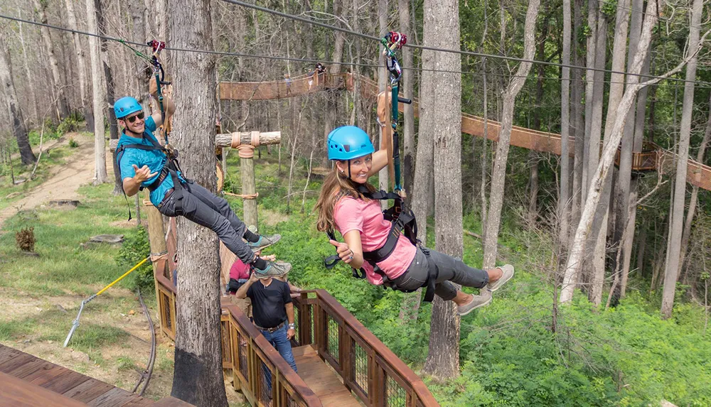 Two people are enjoying a zipline adventure in a wooded area wearing helmets and safety harnesses