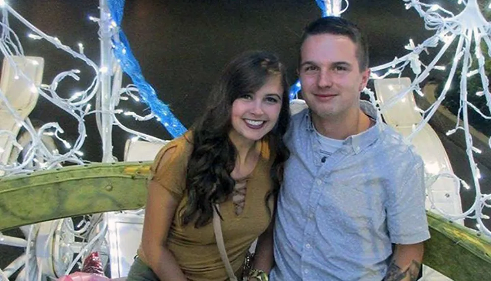 A man and a woman are smiling for a photo in front of a decorative background with white branches and blue lights