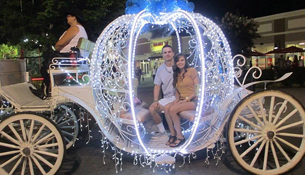 A couple is smiling for the camera while seated in an ornate illuminated carriage reminiscent of Cinderellas pumpkin carriage