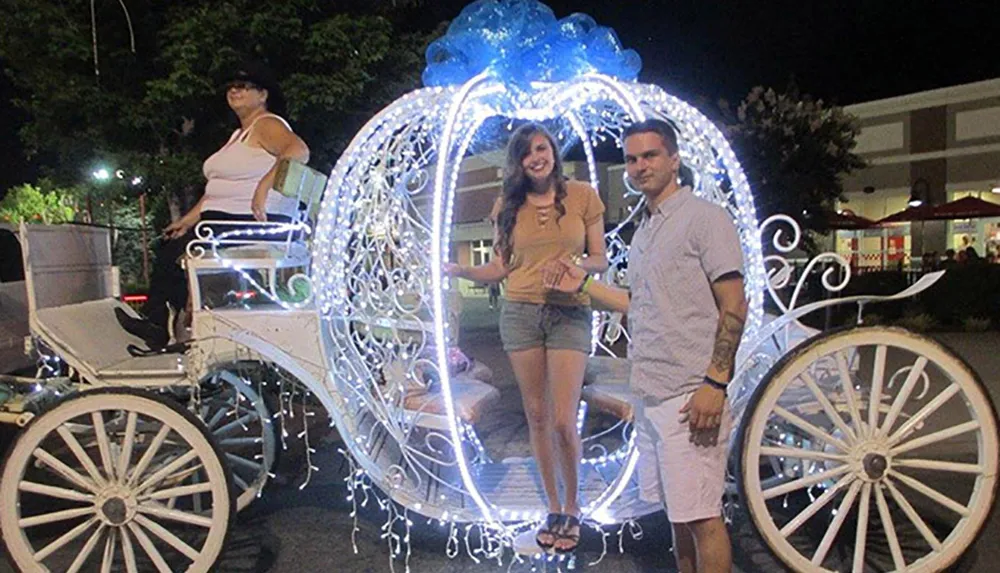 A couple is posing for a photo in front of an ornately decorated illuminated carriage with a driver standing at the back
