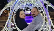 A smiling couple is sitting inside an ornate, light-adorned carriage at night.