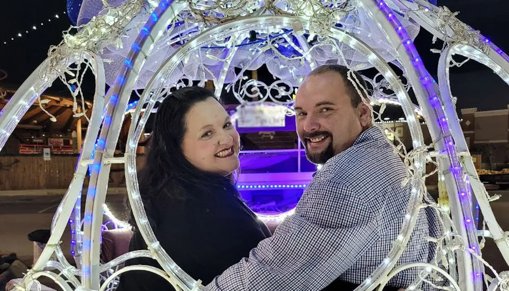 A smiling couple is sitting inside an ornate light-adorned carriage at night