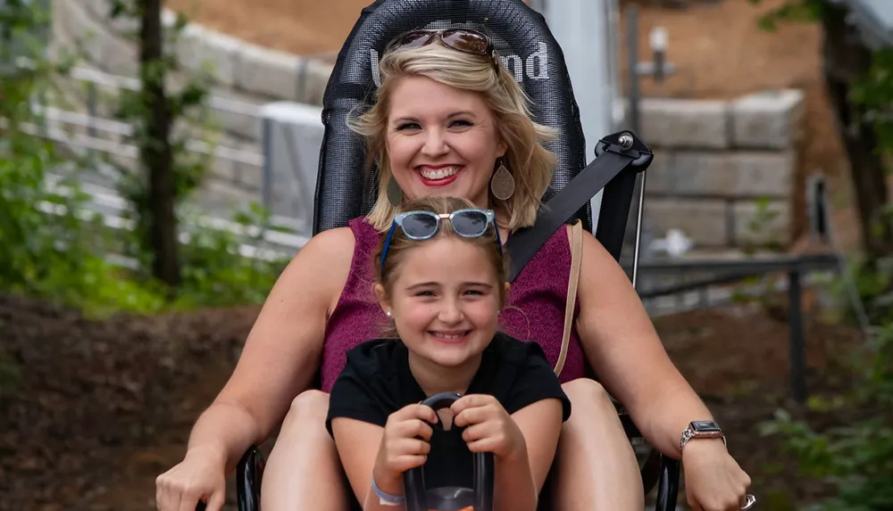 A smiling woman with sunglasses on her head and a young girl in front are enjoying a ride in an outdoor setting both looking joyful and excited