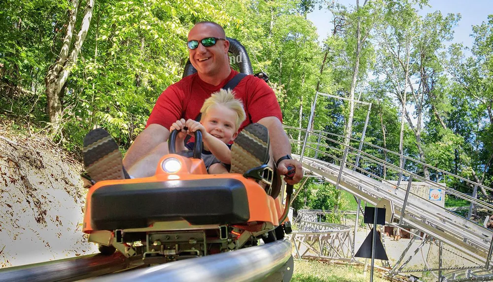 A child and an adult are enjoying a ride on a mountain coaster amidst a wooded area