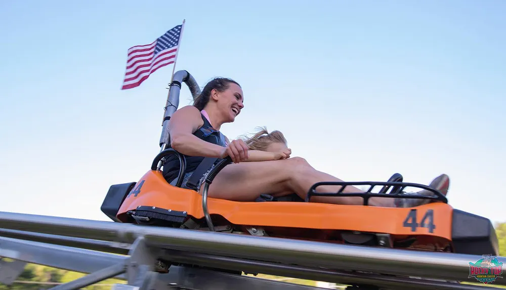 Two people are enjoying a thrilling ride on an orange mountain coaster with an American flag displayed prominently in the background