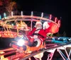 A person dressed as Santa Claus is joyfully waving while riding a small roller coaster at nighttime illuminated by colorful lights