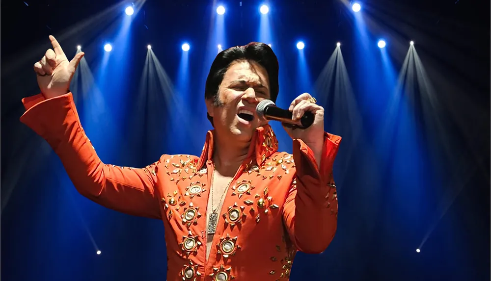 A performer dressed in an embellished red jumpsuit is singing into a microphone under blue stage lights