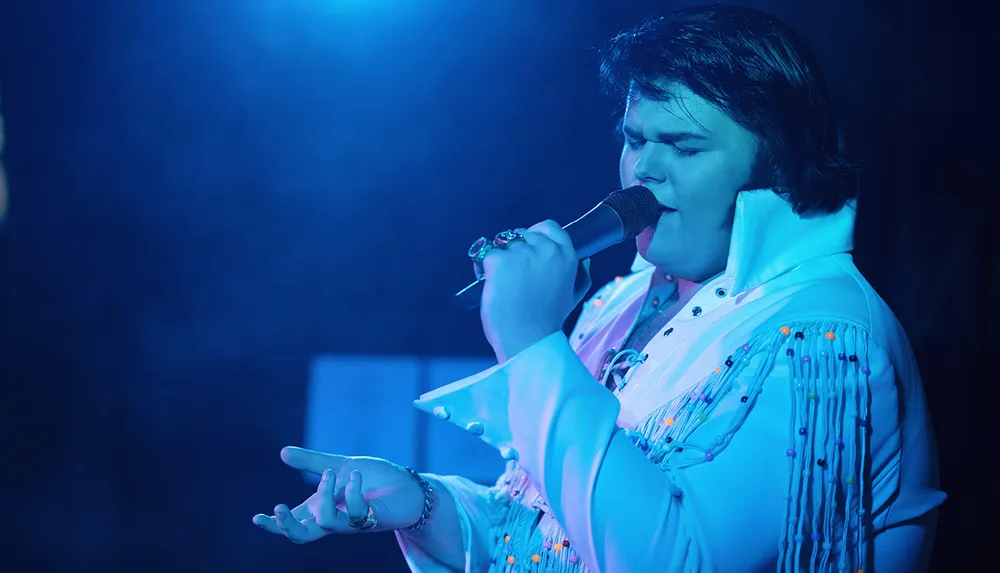 A person in an Elvis-style outfit is passionately singing into a microphone under blue stage lighting