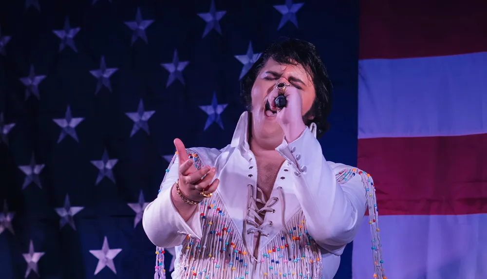 A person dressed in a white bejeweled jumpsuit sings passionately with a microphone against a backdrop of an American flag