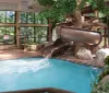 The image shows an indoor swimming pool with a water slide surrounded by a faux-naturalistic decor and large windows that reveal outdoor greenery