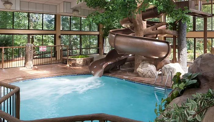 The image shows an indoor swimming pool with a water slide surrounded by a faux-naturalistic decor and large windows that reveal outdoor greenery
