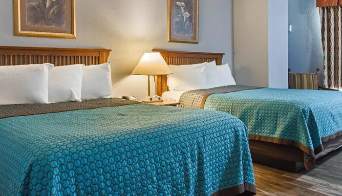 The image shows a neatly arranged hotel room with two beds covered in turquoise bedspreads wooden headboards a shared nightstand with a lamp and tasteful wall art