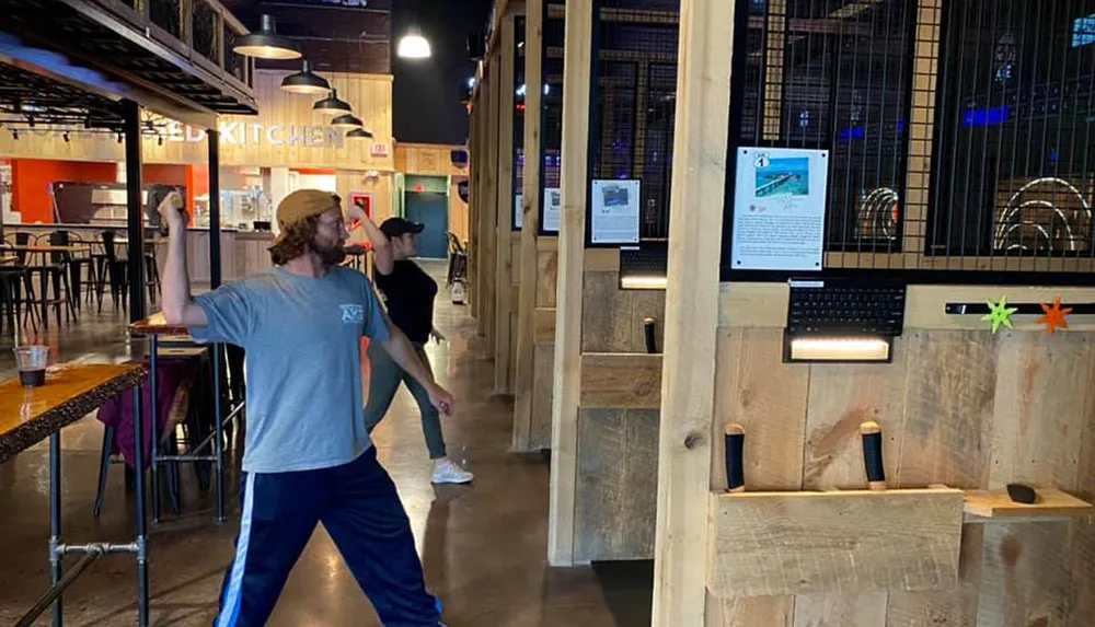 Two people are playing an axe throwing game at an indoor venue with an onlooking food service area in the background
