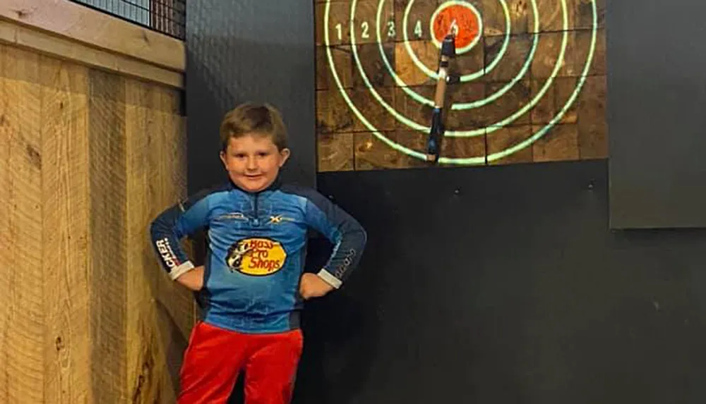 A confident young boy is posing with his hands on his hips in front of a dartboard where a dart has hit the bullseye